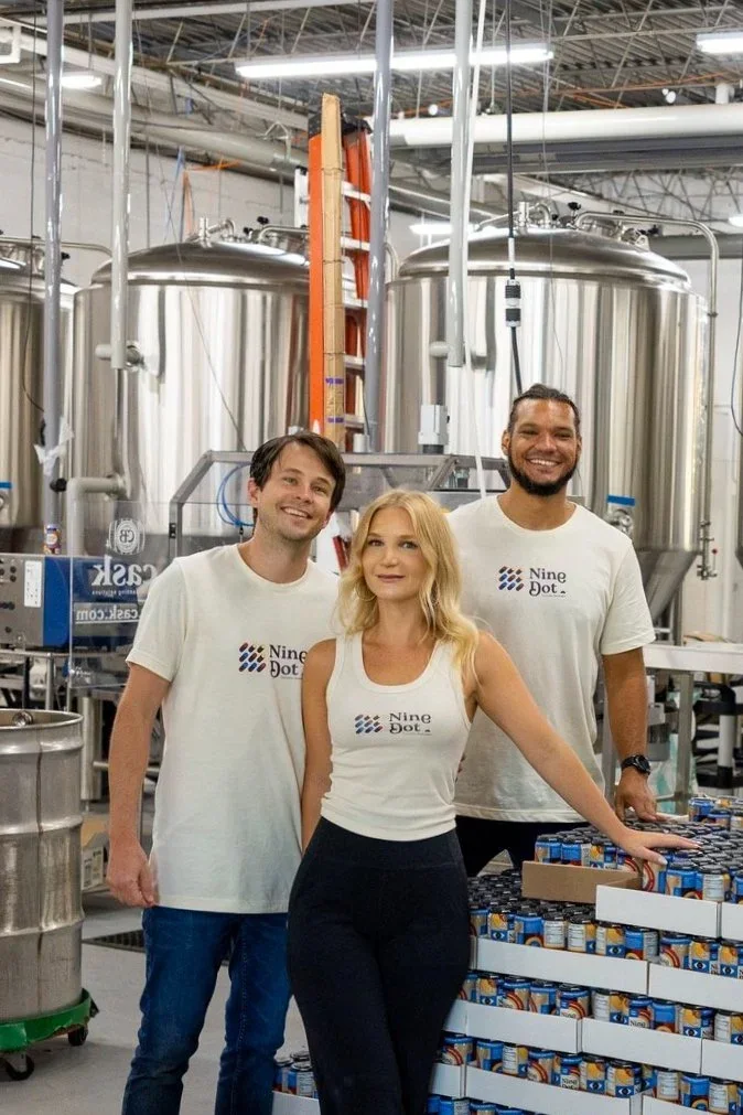 David Spang, Crystal Spang, and a Nine Dot team member at the production facility, in front of stainless brew tanks and pallets of cans
